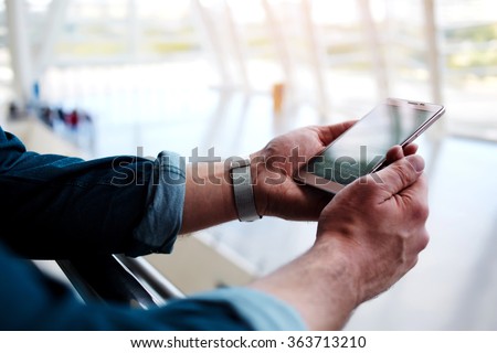 Cropped shot view of a man\'s hands holding cell telephone while standing in shopping center, young male connecting to wireless via mobile phone while waiting for someone in modern airport hall