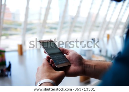 Cropped shot view of man\'s hands orders electronic ticket via mobile phone application during summer trip, young male reading text message on cell telephone while standing in waiting hall of airport