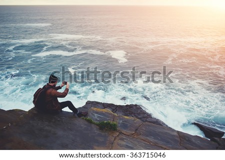 Hipster guy taking pictures of amazing landscape on mobile smart phone digital camera while sitting on a rock near the sea, beautiful sunset light create background with copy space for text message