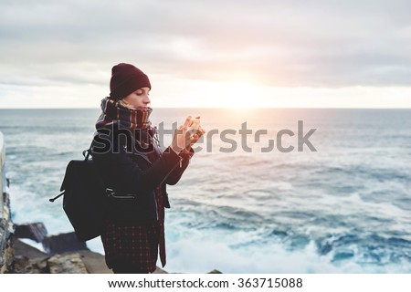 Woman tourist dressed in stylish clothes shoots video of beautiful sea landscape on cell telephone, hipster girl taking photo with mobile phone camera while standing near ocean in cool spring day