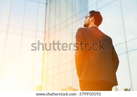 Back view of rich businessman with hands in pockets looking on his skyscraper building while standing outside, young confident male manager dreaming about growth his career during work break outdoors