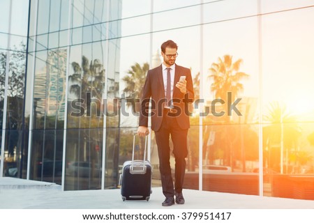 Elegant businessman checking e-mail on mobile phone while walking with suitcase outside airport, experienced male employer using cell telephone while waiting for taxi car outdoors before work travel