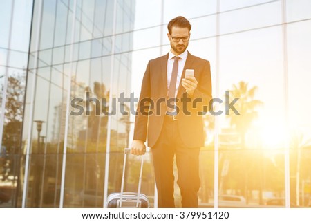 Young male CEO with suitcase reading text message on cell telephone while walking outdoors near building with city reflection, intelligent employer dials the number on smart phone before business trip
