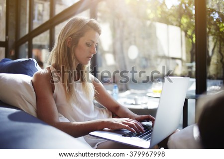 Charming European woman chatting in social network via net-book while sitting in modern interior, beautiful young female searching work via internet on laptop computer during free time in weekend