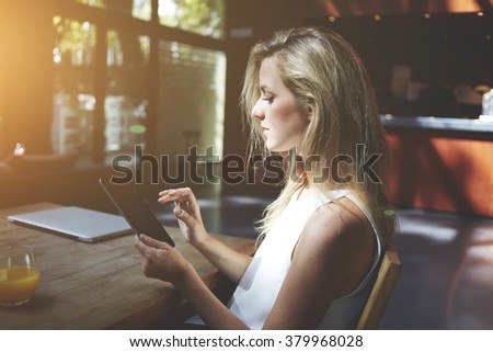 Female searching necessary information in network via touch pad while sitting at the table with closed net-book in co-working cafe, young woman chatting with friends on touch pad while relaxing in bar