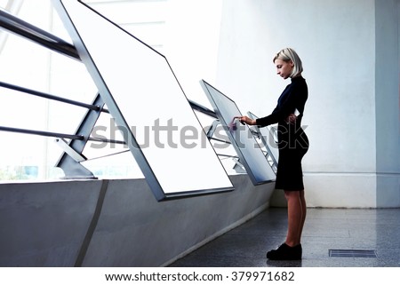 Young businesswoman working on interactive display while standing near another big computer monitor with blank copy space screen for your text message or content, elegant female selecting information