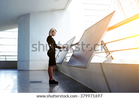 Beautiful female reading advertising about a project on interactive computer display while standing in office interior, businesswoman searching information on high tech modern device