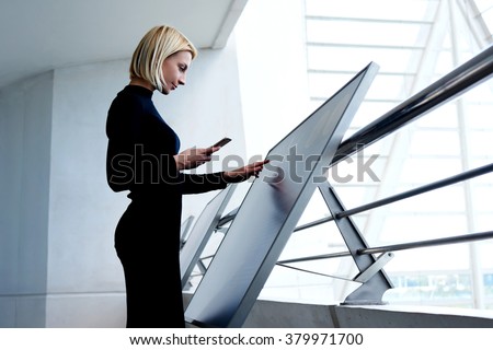 Young intelligent female phoning to partner via mobile phone while selecting information on high tech device, smart businesswoman holding cell telephone while exploring project on interactive monitor