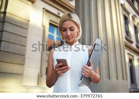 Pretty businesswoman reading news via cell telephone while standing outdoors before presentation on net-book, young female holding laptop computer in hand and writing text message on mobile phone