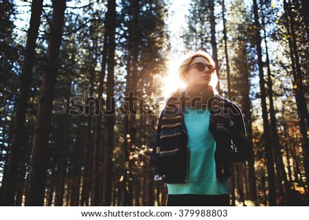 Woman tourist enjoying fresh air while resting after strolling in forest in sunny autumn day, female in trendy sunglasses admires nature while standing near tall trees in the park during fall weekend