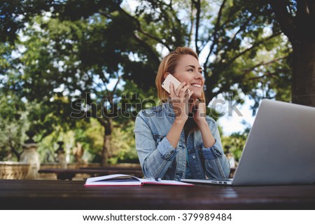 Young happy woman calling with smart phone while taking break between work on laptop computer, smiling female student talking on cell phone while sitting with net-book in park during recreation time