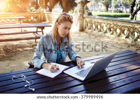 Young female student writes information from portable net-book while prepare for lectures in University campus,hipster girl working on laptop computer while sitting in city park during recreation time