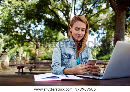 Beautiful woman chatting on mobile phone while sitting with portable laptop computer on campus, young female in good mood reading text message on cell telephone while resting after work on net-book
