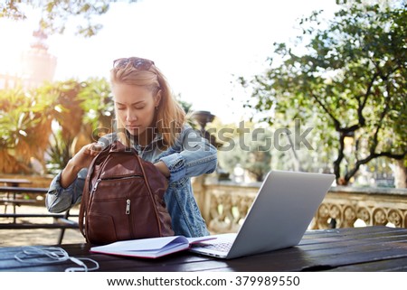Hipster girl looking for something in the bag while sitting outdoors at the table with open net-book, young female searching the pen in rucksack for writing an essay on a notebook from laptop computer