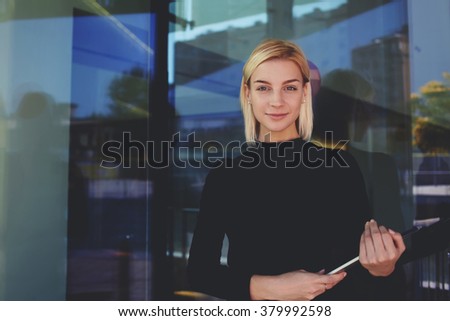 Successful business owner posing with touch pad while standing outside near glass entrance door of office building,confident woman entrepreneur holding digital tablet and look at the camera with smile