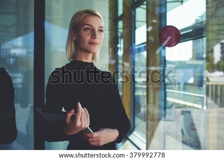 Young female real estate agent with touch pad waiting for tenant while standing in modern building hallway, confident businesswoman holding digital tablet during encounters foreign partners in airport
