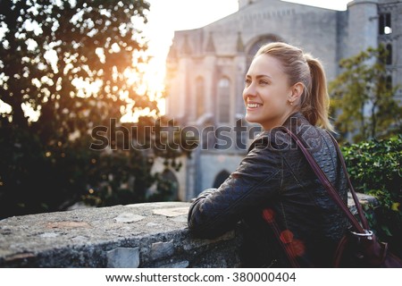 Young smiling woman traveler enjoying town landscape while standing outdoors against big old church, cheerful hipster girl with a rucksack on her back looking at beautiful places from balcony outdoors