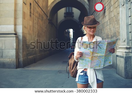 Happy female tourist with backpack on shoulders exploring map while standing in alley near vintage building, trendy woman traveler with smile reading geographical atlas while touring in old city