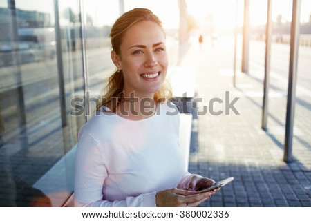 Pretty European woman with beautiful smile holding smart phone while sitting on a bus stop in urban setting, cheerful attractive female using cell telephone while waiting outdoors her friends