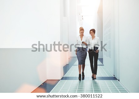 Two confident female leaders reading news on mobile phone about their collaboration while walking together in office, businesswomen watching something on cell telephone while goes in hallway company