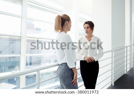 Elegant women entrepreneurs speaking about something personal while standing near office window, two European female managers dressed in formal wear have met in company corridor during work break