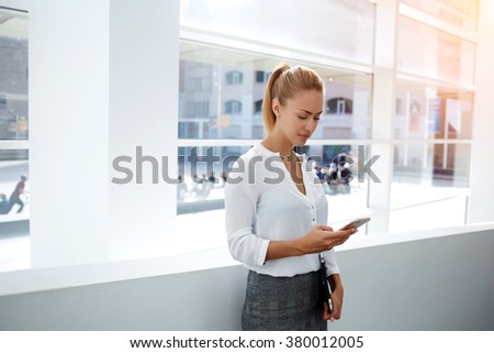 Beautiful young businesswoman dressed in corporate clothing browsing wireless on cell telephone during work break,female financier chatting on her mobile phone while standing in modern office interior