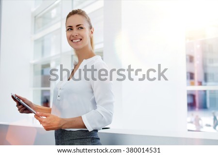 Happy woman manager holding touch pad and thinking about something good while standing in modern office interior,young smart female secretary smiling for someone during work on portable digital tablet