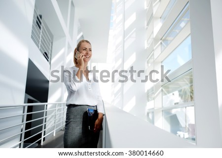Businesswoman with digital tablet in hand talk on mobile phone with partner about successful project presentation, smiling female proud ceo speaking on cell telephone while standing in office interior