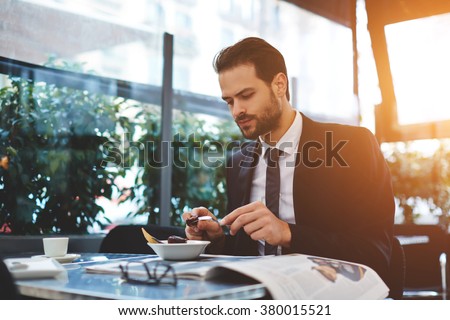 Young confident businessman in luxury suit having breakfast in modern coffee shop during work break, serious male restaurant owner eating tasty dish which was prepared chief cook specially for him
