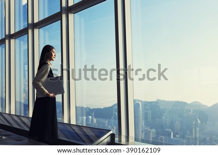Young Asian executive holding laptop computer, while is standing in office interior and looking out of big window with city view. Female manager with net-book in hand ponder about new business project