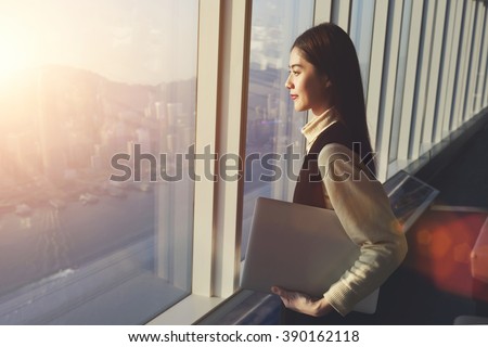 Young woman candidate is holding laptop computer, while standing in office interior and looking out of big window with city view. Female manager with net-book in hand ponder about new business project