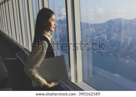 Chinese female successful entrepreneur with laptop computer is standing in modern office interior near skyscraper window with copy space area for your advertising text message or promotional content