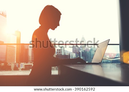 Silhouette of a woman intelligent managing director is keyboarding on laptop computer, while is sitting in modern office interior. Female economist is analyzing the activities of company via net-book