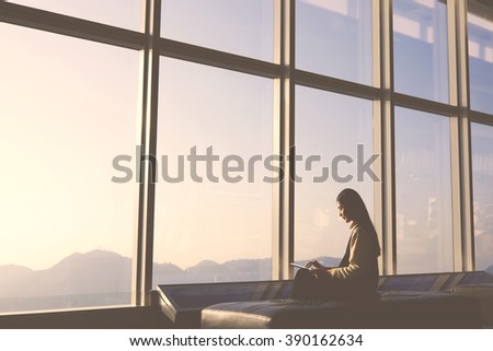 Young female traveler is sitting in modern airport interior and connecting to free wireless for make hotel booking in city of arrival. Silhouette of woman is using digital tablet and waiting her fly