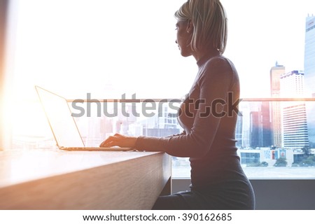 Charming European woman chat in social network via net-book while sitting in modern interior with city scape outside the window, Beautiful young female searching work via internet on laptop computer