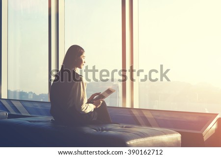 Young Asian hipster girl is holding touch pad, while is sitting in modern interior against window with city view. Businesswoman is waiting her fly in airport and thinking about her awaited vacations