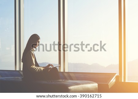 Young Asian female is watching news on digital tablet, while is sitting in airport interior against window with copy space area.Attractive Chinese woman is using portable computer and waiting her fly