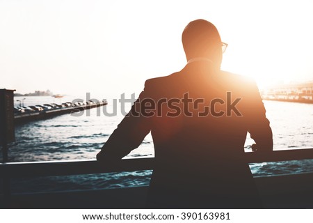 Back view of a young businessman is enjoying view and rest after meeting during his business trip. Silhouette of a male lawyer is resting after work day, while is standing outdoors against sunset