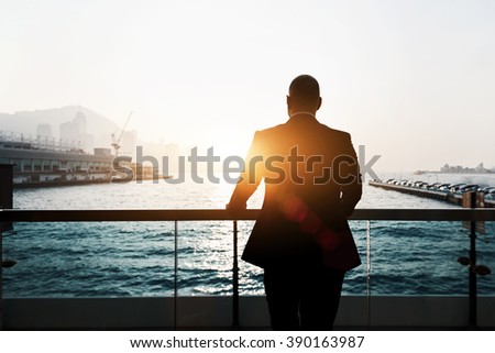 Back view of male successful entrepreneur enjoying evening sunset while standing outdoors in marina port. Silhouette of male skilled and confident businessman is thinking about his future plans