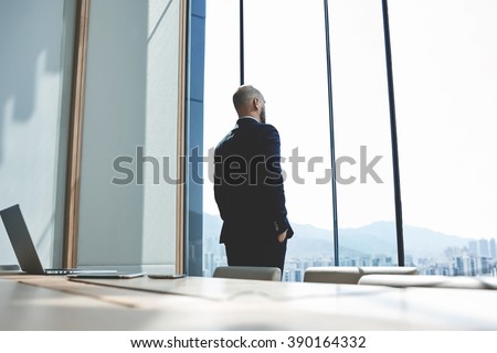 Back view of a successful boss looking through a window, while is standing in his modern office. Young man skilled entrepreneur in rich suit is thinking about meeting with shareholders of the company