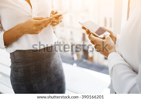 Closely image of a woman financier is reading financial news in web via smart phone, while her partner is typing text message on cell telephone. Two female are using their cellphones during work break