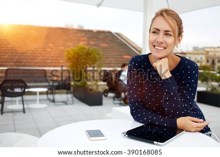 Smiling woman is sitting in comfortable cafe-bar with portable touch pad during her recreation time, cheerful happy female rests after work on digital tablet and waits her friends in cozy coffee shop
