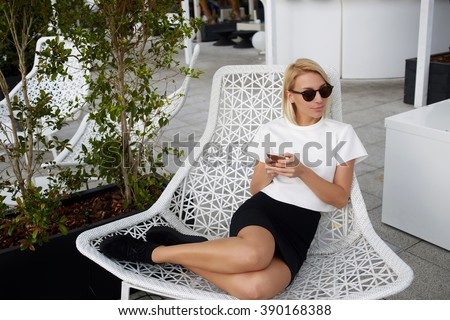Charming blonde female is looking away, while is sitting in comfortable chair with mobile phone in hands. Hipster girl in fashionable clothes is using cell telephone during rest in modern coffee shop