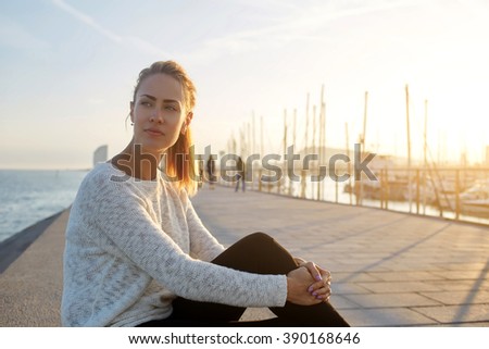 Beautiful thoughtful female tourist enjoys good summer evening and recreation time while sits on pier near sea, cute hipster girl dreams about something and relaxes in the fresh air in her weekend