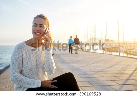 Happy hipster girl having pleasant mobile phone conversation with boyfriend while is sitting near sea in summer evening, smiling female talks on cell telephone and relaxes outdoors during free time