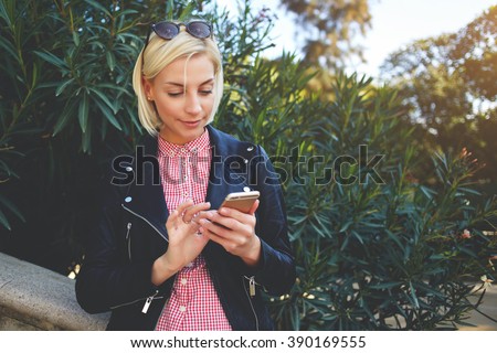 Young hipster girl with trendy look chatting on smart phone while resting in park in spring day, stylish female student searching information in network while sitting at the campus during class break