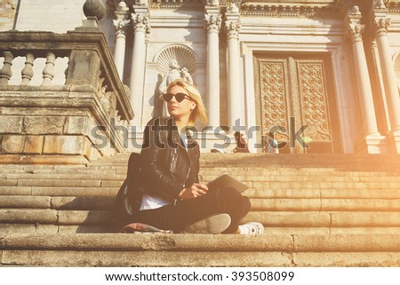 Hipster girl in sunglasses is looking away, while is sitting with touch pad against old architectural building. Young woman tourist is holding digital tablet, while is resting after tour in museum