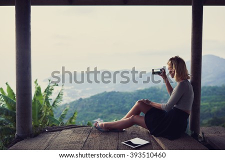 Young woman wanderer is shooting video on her mobile phone, while is sitting against subtropical landscape and sky background with copy space for your advertising text message or promotional content