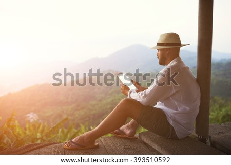 Young man traveler is holding digital tablet with empty screen background with copy space for your advertising content. Male tourist is using touch pad, while is sitting against amazing jungle scenery