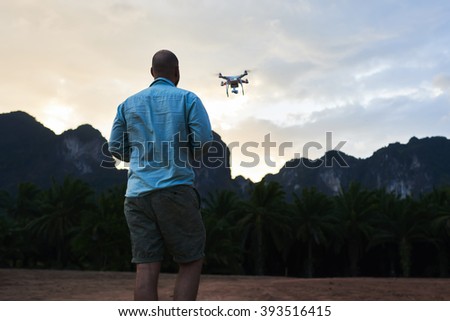Back view of man wanderer is shooting video with radio controlled drone, against silhouette of rock mountains at sunset. Male web author is taking photos on flying quadcopter during trip in Asia